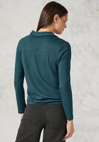 Teal-langärmliges Oberteil mit Kragen, weicher Struktur und elastischem Bund, kombiniert mit dunkelgrauen Hosen, von hinten gezeigt.