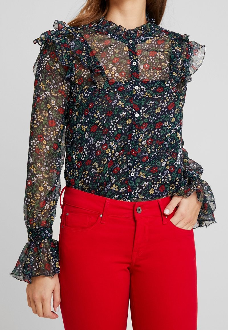 Blouse à manches longues à motifs floraux en tissu noir transparent, ornée de fleurs multicolores, col à volants et fermeture par boutons, associée à un jean rouge.