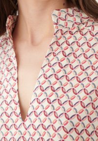 Chemise à col présentant un motif répétitif de boucles entrelacées dans des tons de rouge, rose et bleu sur un fond crème. Texture de tissu lisse.