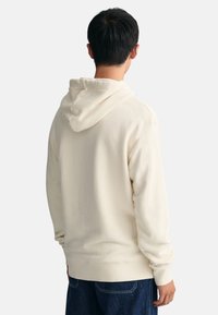 Kremowa bluza z kapturem wykonana z miękkiego materiału, z regulowanym kapturem na sznurki i ściągaczami przy mankietach. Widok z tyłu ukazuje luźny fason i prosty design.