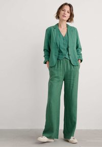 Vihreä pellavapuku, johon kuuluu istuva pitkähihaista bleiseri, napitettava liivi ja leveälahkeiset housut. Rakenteellinen kangas, joka on rento ja mukava.