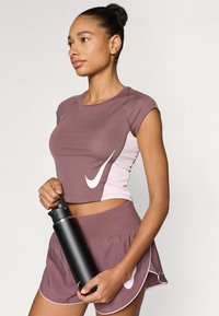 Αθλητικό σύνολο που περιλαμβάνει ένα εφαρμοστό, κοντομάνικο τοπ σε αποχρώσεις μωβ με ροζ λεπτομέρειες και ένα εμφανές λευκό λογότυπο, συνδυασμένο με αντίστοιχα σορτς και ένα μαύρο παγούρι νερού.