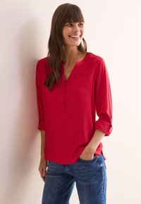 Blusa rossa a maniche lunghe con scollatura a V e patta con bottoni. Tessuto morbido, vestibilità comoda e maniche arrotolate. Abbinata a jeans in denim blu.
