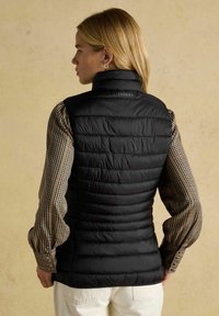 Fekete steppelt mellény magas gallérral és Joules logóval, kockás ing fölött viselve; vízszintes varrásokkal és enyhén fényes felülettel rendelkezik.