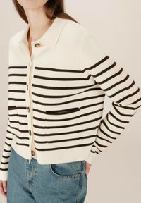 Gilet rayé en crème et marron avec un col, des boutons sur le devant et deux poches avant. Matériau en tricot doux avec une coupe décontractée.