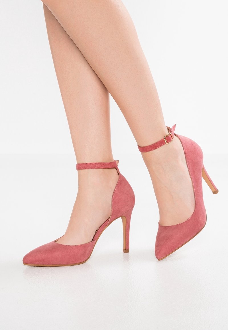 Jambes portant des chaussures à talons hauts en daim rose à bout pointu, avec des brides cheville et une boucle dorée, debout sur un sol blanc réfléchissant.