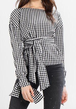 Kvinde iført en sort-hvid gingham langærmet top med taljebånd kombineret med sorte jeans med små pynt.