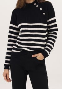 Femme portant un pull en tricot à rayures horizontales noires et blanches avec quatre boutons argentés près de l'épaule, assorti à un pantalon noir.