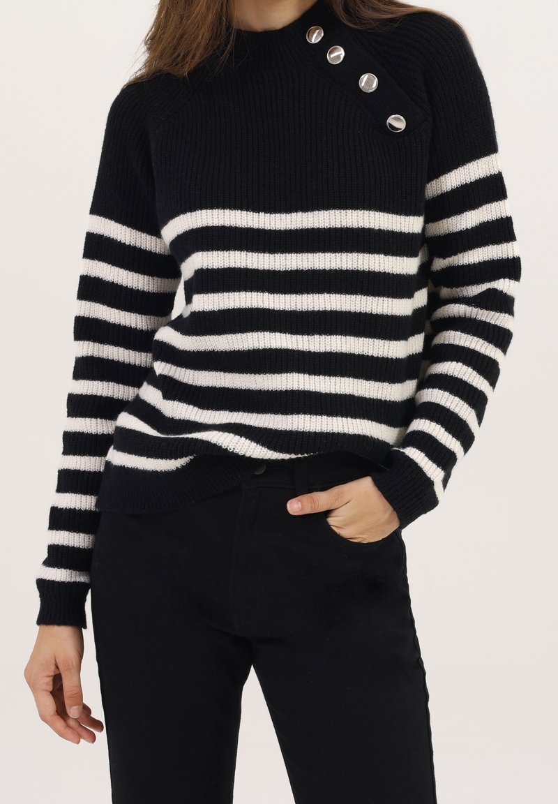 Femme portant un pull en tricot à rayures horizontales noires et blanches avec quatre boutons argentés près de l'épaule, assorti à un pantalon noir.