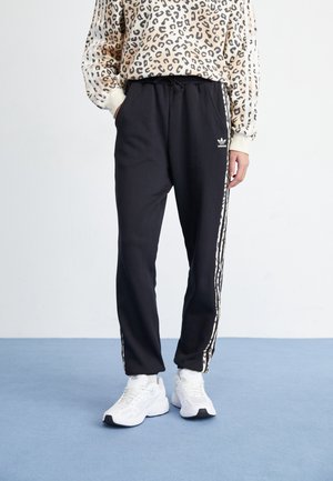 Pantalones deportivos negros con rayas blancas laterales, con cintura elástica y logo. Combinados con una camiseta de manga larga estampada y zapatillas blancas.