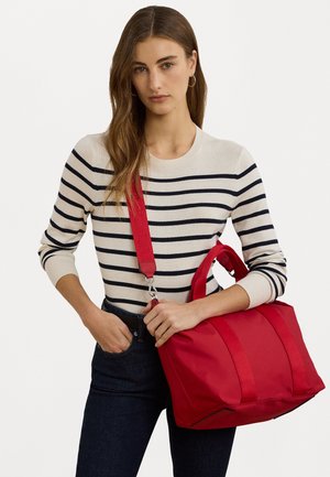 Jonge vrouw met lang bruin haar, gekleed in een wit en marineblauw gestreepte trui en donkere jeans, die een rode schoudertas vasthoudt.