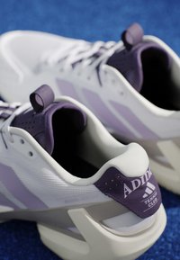 Paire de chaussures de tennis Adidas blanches et violettes posées sur une surface bleue, montrant le logo et un design à lacets avec col rembourré.