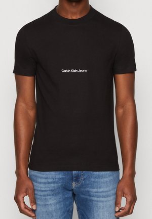 Mand iført en tætsiddende sort Calvin Klein Jeans t-shirt med hvidt logo i midten af brystet og blå denimjeans.