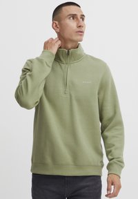 Sudadera de forro polar verde con cuello de media cremallera, que presenta un pequeño logo en el pecho, puños acanalados y un diseño de corte relajado.