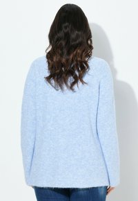 Frau mit welligem, dunklem Haar, die einen hellblauen, flauschigen Pullover und dunkelblaue Jeans trägt, vor einem schlichten weißen Hintergrund stehend.