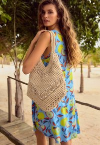 Sac en crochet beige avec un motif en chevrons, orné d'accents en perles. Le modèle porte une robe bleue avec un imprimé de fruits coloré.