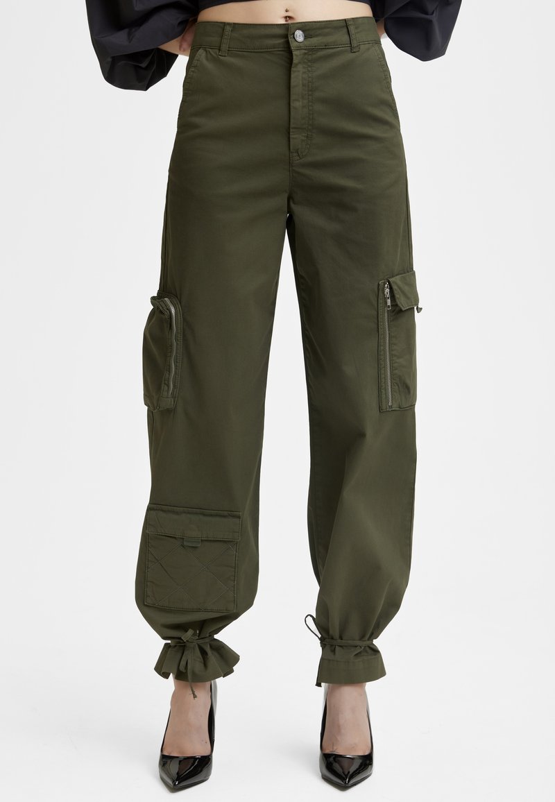 Pantalon cargo vert olive avec plusieurs poches à zip et à rabat, ourlets à cordon, porté avec des talons noirs à bout pointu.