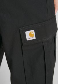 Černé cargo kalhoty vyrobené z texturované tkaniny, s kapsou na klopu a výraznou žluto-bílou nášivkou Carhartt.