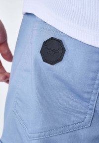 Tasca di pantaloni blu chiaro con toppa a logo ottagonale nera, mano vicino alla tasca e tessuto a coste bianco visibile sul bordo superiore.