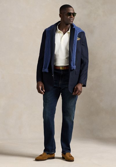 Marineblå blazer over en hvit polo, blå strikkegenser lagt over skuldrene, mørke jeans, brune loafers og solbriller. Enkelt, klassisk design.