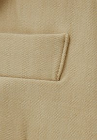 Beżowy materiał z subtelną diagonalną teksturą posiada jedną, skośną kieszeń. Szwy są widoczne wzdłuż krawędzi.