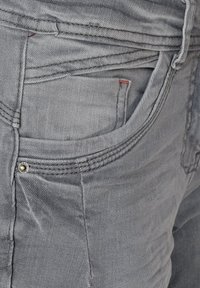 Jeans de mezclilla gris con una superficie texturizada, que presentan un pequeño bolsillo, costuras visibles y un acento de botón de latón.