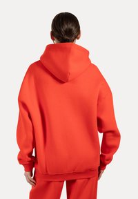 Oranje hoodie met een relaxte pasvorm, ribgebreide manchetten en zoom, en zachte katoen. De achterkant toont de plaatsing van de capuchon en de naaddetails.
