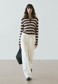 Gestreiftes Langarmshirt in Beige und Braun, kombiniert mit weiten weißen Hosen. Das Modell trägt eine strukturierte schwarze Handtasche und schwarze Schuhe.