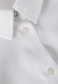 Bílá košile s knoflíky vyrobená z hladké tkaniny, s špičatým límcem a průhlednými knoflíky se subtilním logem na výstřihu.