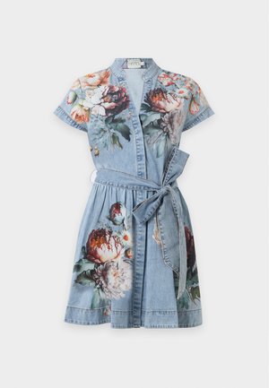 Ung kvinde iført en kortærmet, bæltet denimkjole med blomsterprint, stående og poserende med beige højhælede sko mod en ensfarvet baggrund.