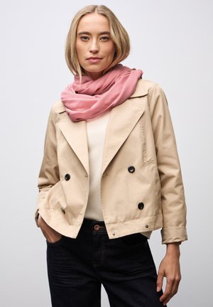 Veste beige à double boutonnage avec de grands boutons noirs, portée sur un pull crème, associée à une écharpe rose. Un jean en denim sombre complète le look.