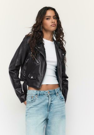 Junge Frau mit langem, welligem Haar, die eine schwarze Lederjacke, ein weißes Croptop, hellblaue Jeans und Creolen trägt und vor einer schlichten Wand posiert.