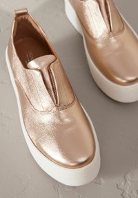 Chaussures slip-on en rose gold métallique avec une texture lisse et une semelle plate blanche. Comprend un détail de couture à l'avant et une languette.