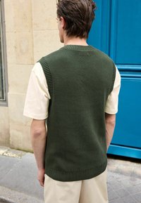 Homme portant un gilet en tricot vert foncé par-dessus une chemise à manches courtes crème, debout en extérieur près d'un mur beige et d'une porte bleue.