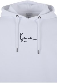 Biela mikina s kapucňou vyrobená z mäkkej tkaniny, na hrudi s čiernym vyšívaným logom. Má šnúrku s kovovými striebornými koncami.