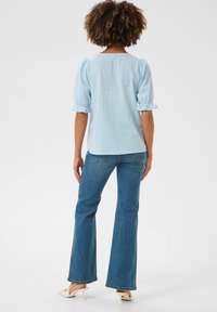 Világoskék, rövid ujjú felső fodros mandzsettákkal, bő szabású kék farmernadrággal párosítva. A modellt hátulról látjuk, egy egyszínű háttér előtt.