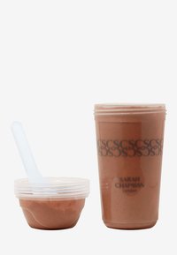 Zwei transparente Behälter: eine kleine Schüssel mit einem durchsichtigen Löffel und eine größere Tasse, gefüllt mit einer dicken, braunen Substanz, die mit "Sarah Chapman London" beschriftet ist.