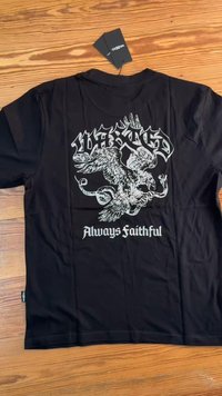 Fekete pamut póló, amelyen egy fehér sárkány-szerű lény grafikája látható, alatta pedig a "Mindig Hű" felirat, címkével ellátva.