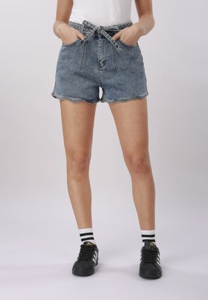 Kobieta w wysokich niebieskich krótkich spodenkach dżinsowych z przewiązanym paskiem, białych skarpetach z czarnymi paskami oraz czarnych tenisówkach Adidas, z rękami w kieszeniach.