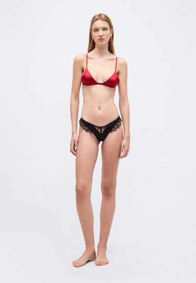 Νεαρή γυναίκα στέκεται ξυπόλητη φορώντας κόκκινο σατέν σλιπ με λεπτές τιράντες και μαύρο εσώρουχο με δαντέλα, μπροστά σε απλό φωτεινό φόντο.