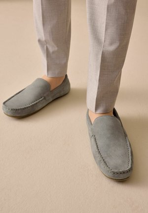 Osoba stojící na béžové podlaze, obutá v nazouvacích mokasínech z šedé semiše a sladěných světle šedých kalhotách.