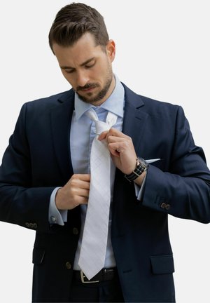 Cravate blanche à motifs en texture de diamant, portée avec une chemise bleu clair et un costume sombre. Un homme ajuste sa cravate, mettant en valeur une montre à son poignet.