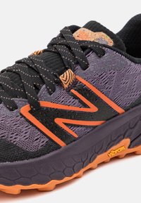 Scarpa sportiva viola e nera con tomaia testurizzata, accenti arancioni e logo Vibram sulla suola. Presenta lacci e un design imbottito.