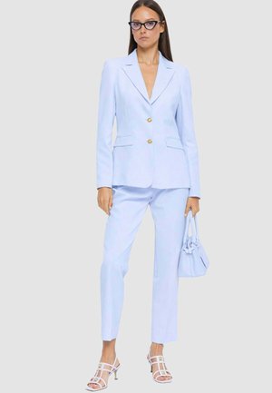 Donna con completo azzurro chiaro su misura con bottoni dorati, tacchi bianchi con cinturini, occhiali neri, che tiene una borsa blu coordinata su sfondo uniforme.