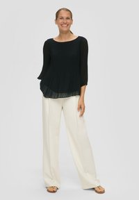 Must mustriga tops, millel on 3/4-sleeved varrukad ja ümmargune kaelus, on kombineeritud kreemikatest laiematest pükstest. Mudel kannab heledaid sandaale. Lihtne taust.