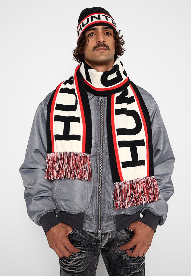 Czarny i biały gruby szal robiony na drutach z czerwonymi i czarnymi akcentami, z frędzlami na końcach i dużymi literami na całej powierzchni. W komplecie z pasującą czapką.