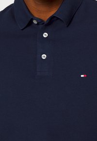 Camisa polo azul marino con cuello abotonado, que presenta una tela texturizada y un pequeño logo en rojo, blanco y azul en el lado izquierdo del pecho.