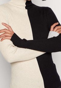 Rippstrick-Turtleneck-Pullover in einem Farbschnitt-Design, mit Creme auf der einen Seite und Schwarz auf der anderen; figurbetonter Stil mit langen Ärmeln.