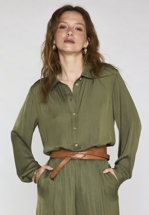 Donna con capelli lunghi e mossi che indossa una camicetta verde oliva con bottoni e pantaloni abbinati, accessoriata con cintura marrone, orecchini a cerchio dorati e braccialetti.