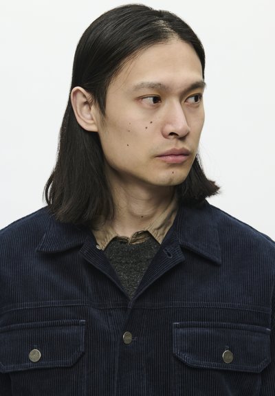 Námořnická kordový kabát s dvěma předními kapsami, světle hnědým límcem a šedým svetr pod ním; obsahuje kovové knoflíky a strukturovanou tkaninu.